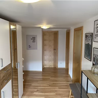 Ein heller Flur mit Holzboden und modernen Möbeln. An den Wänden hängen verschiedene Bilder und ein Spiegel reflektiert den Raum.