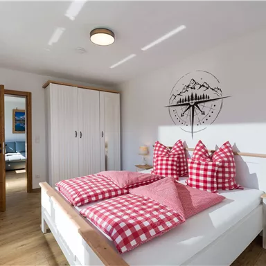 A cozy bedroom with a large bed and checked pillows. The room has a bright, modern look with wood details and a wall decor of mountains.