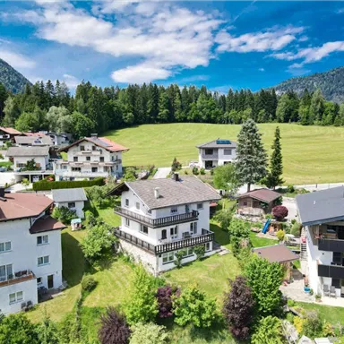 A picturesque landscape with several houses amidst trees and meadows. In the background, gentle hills and a blue sky can be seen.