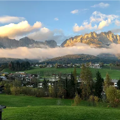 Eine malerische Landschaft mit Bergen und Wolken. Im Vordergrund sind grüne Wiesen und einige Häuser zu sehen.