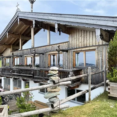 Ein traditionelles Holzhaus in den Bergen mit großen Fenstern und einem Balkon. Im Vordergrund sind einige Pflanzen und ein Steinhaufen zu sehen.