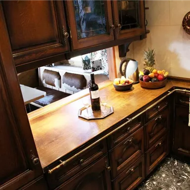 A cozy kitchen with dark wooden cabinets and a wooden countertop. On the counter, there is a bottle of wine and a bowl of fresh fruit.