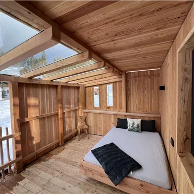 A cozy wooden house with a bed and large windows. Outside you can see snow and trees.
