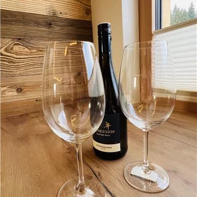 Two wine glasses are on a wooden table next to a bottle of wine. The windows provide a view outside.