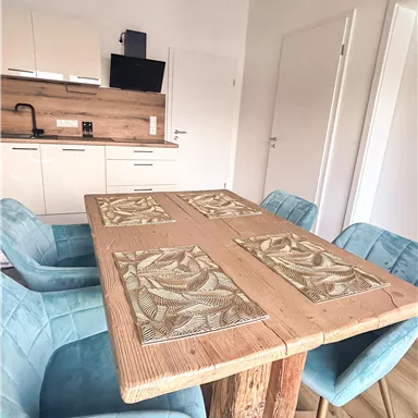 An inviting dining area with a rustic wooden table and blue chairs. The modern kitchen in the background complements the simple, bright design.