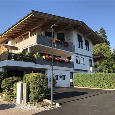 Ein modernes Haus mit einem Balkon und bunten Blumen. Umgeben von grünen Sträuchern und Bäumen.