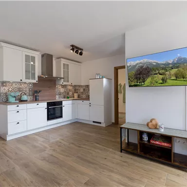 A modern kitchen with white cabinets and wooden flooring. A TV on the wall displays a landscape with mountains.