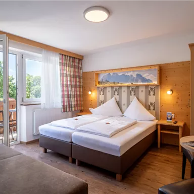 A cozy hotel room with a double bed and wood paneling.
Through the window, view of a green landscape and a balcony.