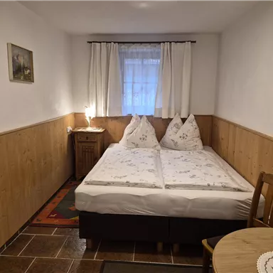 A cozy bedroom with a double bed and two pillows. There is a small table and warm wooden paneling on the walls.