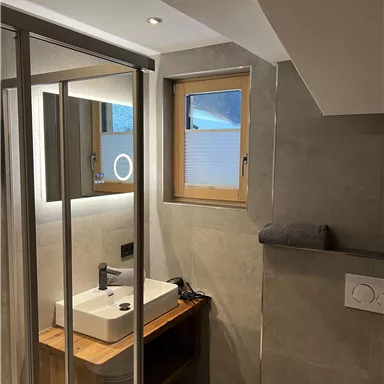 A modern bathroom with a glass shower and a stylish sink. The walls are in a gray concrete look, and a window provides daylight.