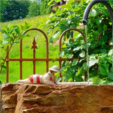 Eine kleine Figur in Badebekleidung liegt entspannt auf einem Stein. Im Hintergrund sprudelt Wasser aus einem Rohr, umgeben von grünen Pflanzen.
