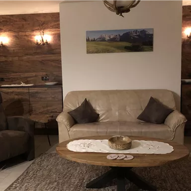 A cozy living room with a beige sofa and two armchairs. The wooden table in the middle is decorated, and the walls are designed with wooden panels.
