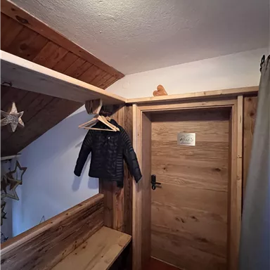 A cozy hallway with a wooden bench and a coat hook. A jacket hangs on the wall, and a wooden door leads to another room.
