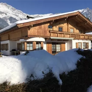 A beautiful chalet in the snow with mountains in the background. The veranda and windows are made of wood and radiate coziness.