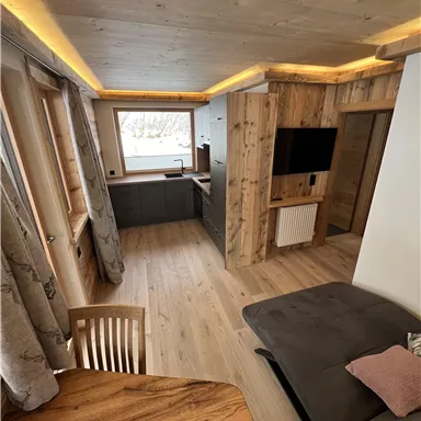 A modern living room with wooden paneling and large windows. The space is cozy furnished with a dining table and a sofa corner.