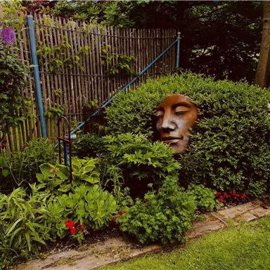 Ein wunderschöner Garten mit üppigem Grün und bunten Blumen. In der Hecke ist eine Skulptur eines Gesichts eingebettet.