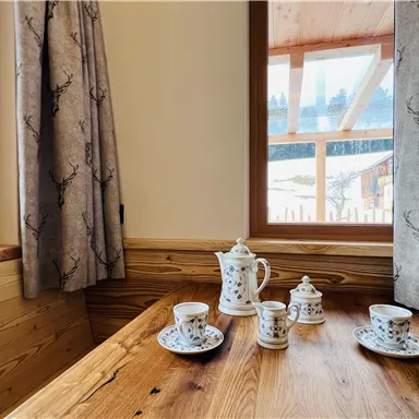 A cozy wooden table with a teapot and porcelain cups. Through the window, you can see a tranquil landscape.