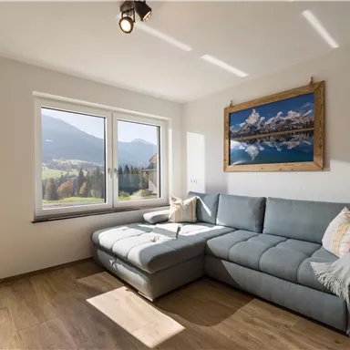 A modern living room with a gray sofa and a beautiful view of the mountains. The light falls through the large windows, creating a bright, inviting atmosphere.