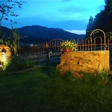 Ein idyllischer Garten bei Dämmerung mit einem großen Kopfskulptur und einem Stein. Im Hintergrund sind sanfte Hügel und eine geschlossene Gartenumzäunung zu sehen.
