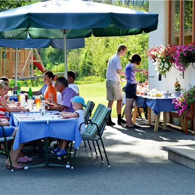 An inviting outdoor area with a table, surrounded by guests. In the background, a person is preparing a buffet while the sun shines.