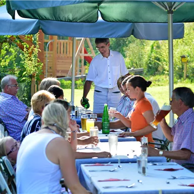 A group of people is sitting at a laid table outdoors. A waiter is serving drinks in a beautiful, green setting.