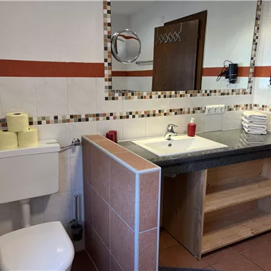 A modern bathroom with a sink, mirror, and shelves. Toilet paper rolls and red candles are decoratively arranged.