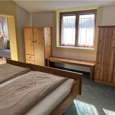 A bright bedroom with a double bed and wooden wardrobes. The room has large windows that let in natural light.