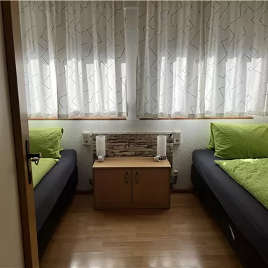 A simple room with two single beds and bright curtains. In the middle, there is a small table with two lamps.