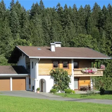 Ein gemütliches Haus in einer idyllischen Lage, umgeben von grünen Wäldern. Der große Garten und die Blumen am Balkon verleihen dem Anwesen einen einladenden Eindruck.