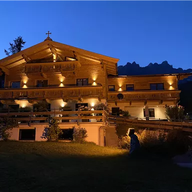 Ein gemütliches Holzhaus, das bei Nacht beleuchtet ist. Im Hintergrund sieht man Berge und einen klaren Abendhimmel.
