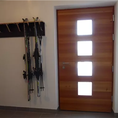 Ein moderner Eingangsbereich mit einer Holzpantentür und drei Fenstern. An der Wand hängen Skistöcke, bereit für den nächsten Ausflug.