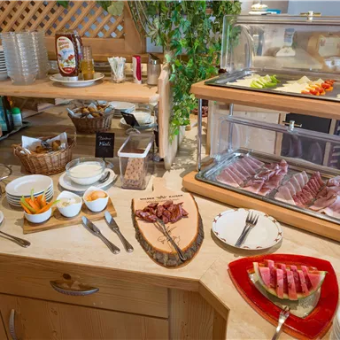 A buffet with various dishes, including cold cuts, cheese, and fruit. Plates and glasses are additionally placed on the table.
