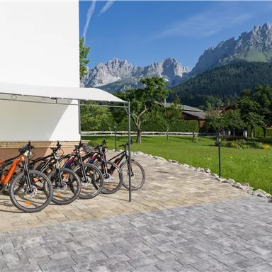 A group of bicycles stands under a canopy on a paved square. In the background, mountains and a green meadow can be seen.