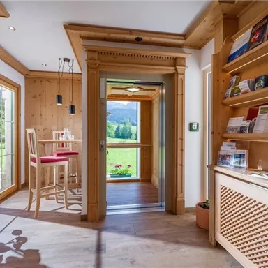 A cozy reception area with wooden paneling and light furniture. A green landscape is visible through the windows.