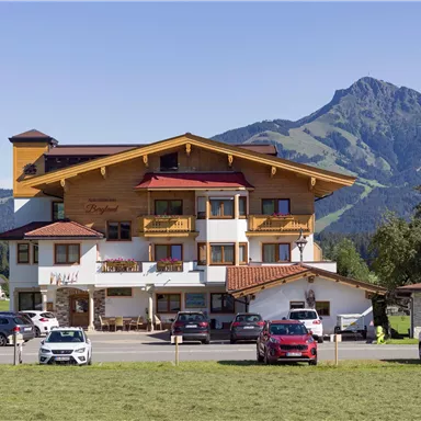A charming hotel with wooden elements, surrounded by green meadows and mountains. The sky is clear and sunny, creating an inviting atmosphere.