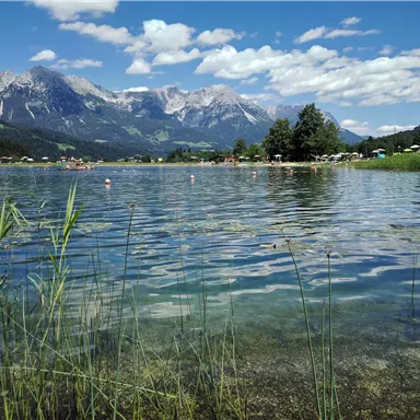 Ein klarer See umgeben von Wiesen und Bergen. Der Himmel ist blau und mit einigen Wolken versehen.