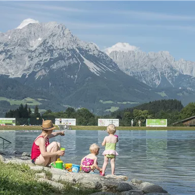 A mother sits by the shore of a lake and plays with two small children. In the background, majestic mountains rise under a blue sky.