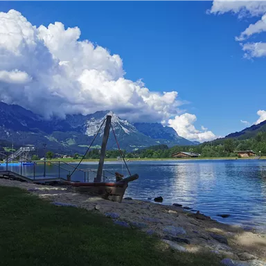 Ein ruhiger See umgeben von Bergen und grünen Wiesen. Die Wolken sind groß und weiß vor einem klaren blauen Himmel.