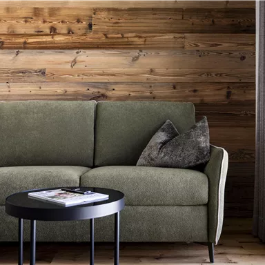 A modern living room with a comfortable, gray-green sofa and a small black table. The wooden wall combined with curtains creates a cozy atmosphere.