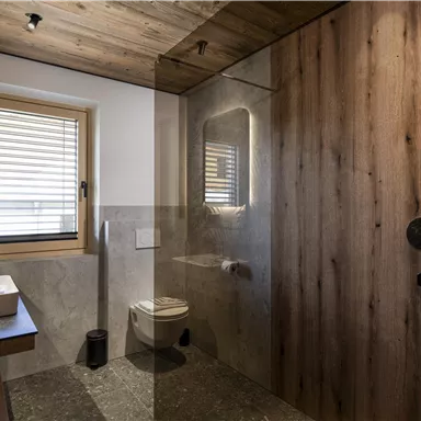 A modern bathroom with wooden paneling and gray tiles. It includes a glass shower, a sink, and a small window.