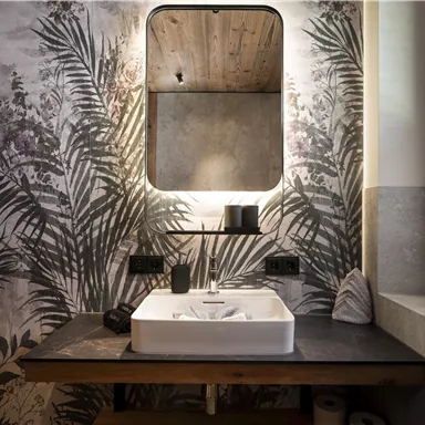 A modern bathroom with a large mirror and a sink on a wooden table.
Walls with a tropical plant motif create a relaxed atmosphere.