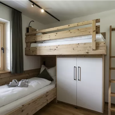 A modern bedroom with a wooden bunk bed and a single bed. The walls are light and there is a large window with a view of the surroundings.