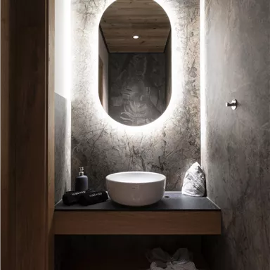 A modern bathroom with elegant lighting and a round mirror. A minimalist wooden vanity top with a white basin and fluffy slippers underneath.