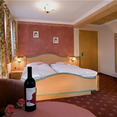 A cozy bedroom with a double bed and warm color scheme. On a table, there is a bottle of wine and a rose.