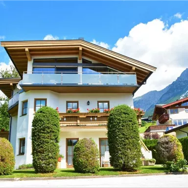 Ein modernes Haus mit einem Balkon und bunten Blumen. Im Hintergrund sind grüne Berge und ein blauer Himmel zu sehen.