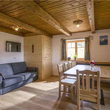 Ein gemütlicher Wohnraum mit Holzdecke und einem großen Esstisch. Das Zimmer ist freundlich und einladend mit einem Sofa und Fenstern, die für viel Licht sorgen.