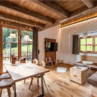 Das Wohnzimmer des Almchalet Hausruck mit einem großen Esstisch und Holzdekor. Durch die Fenster ist die grüne Landschaft sichtbar, die viel Licht hereinlässt.