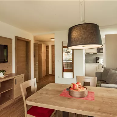 A cozy living space with a dining table, chairs, and a modern kitchen. The room is brightly and friendly designed, with wooden furniture and a large lampshade.