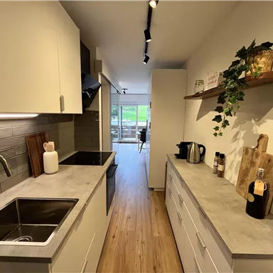 A modern kitchen with light cabinets and a gray countertop. An outdoor area is visible through the window in the background.