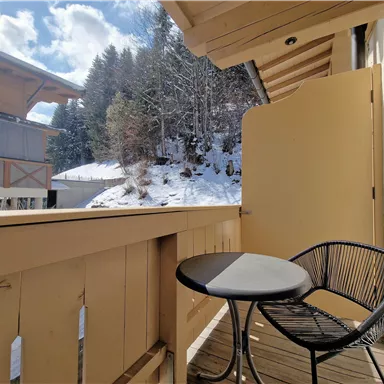 A balcony with a small table and a chair, surrounded by snow-covered trees. The sky is clear and blue, providing a beautiful view.